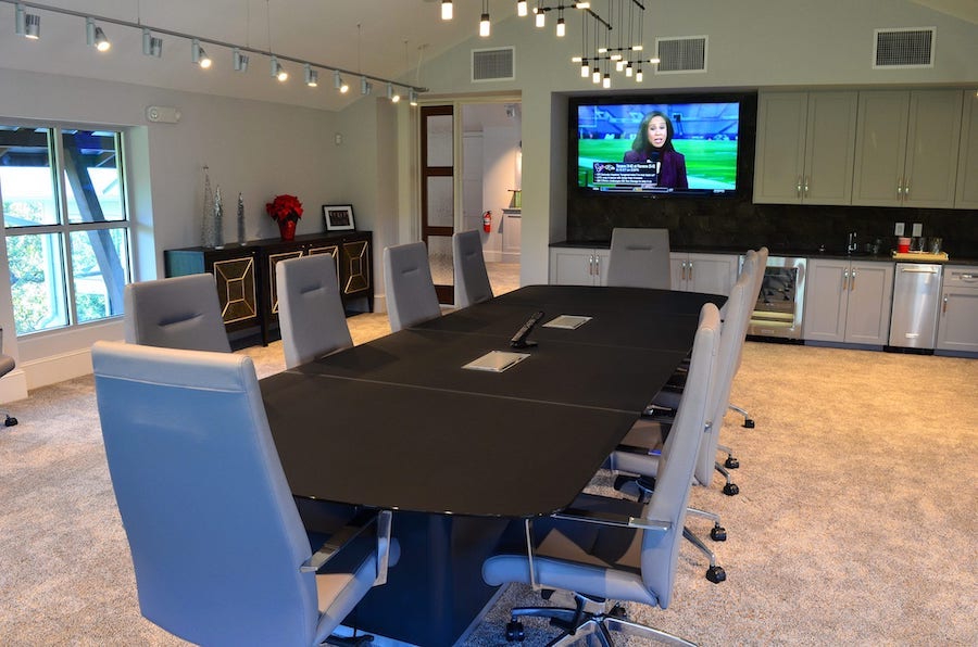 large conference table with tabletop plugs located in front of a wall-mounted TV tuned into a sportscast.